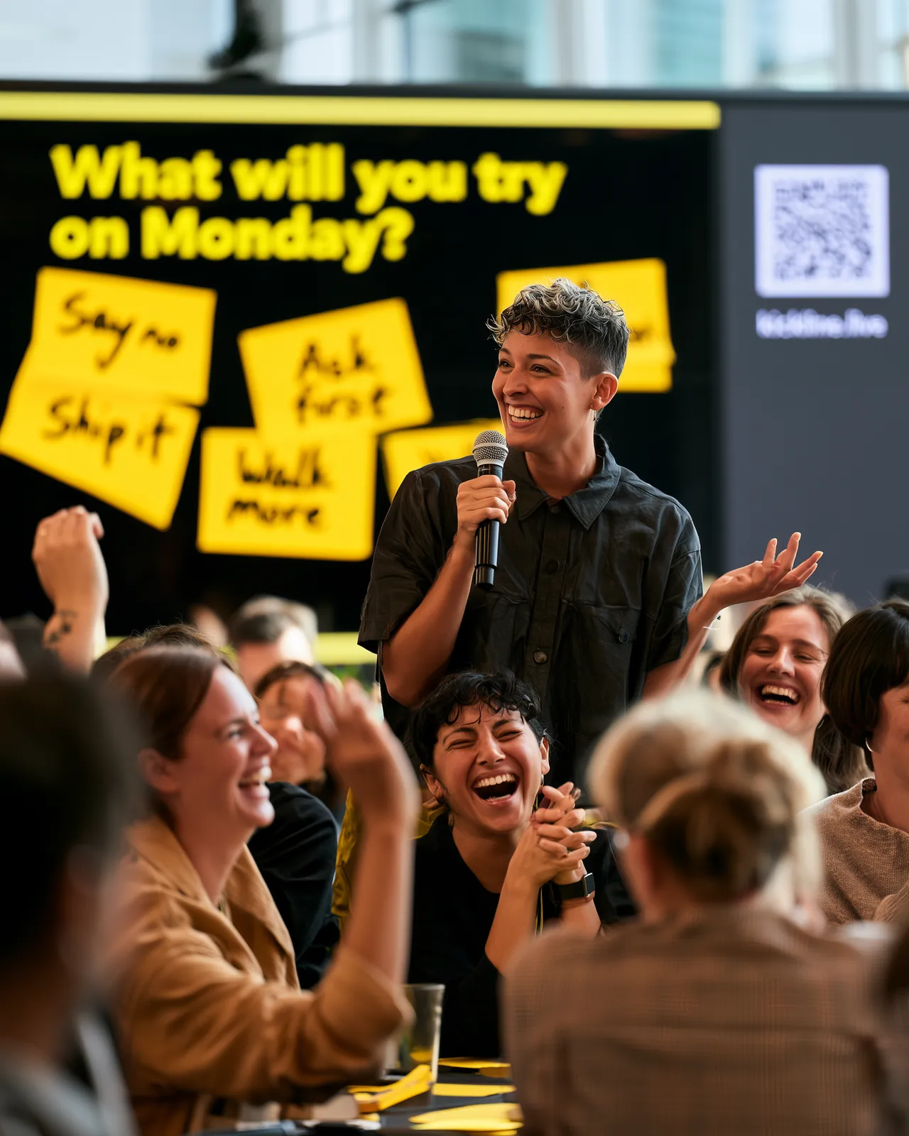 A facilitator laughs with a room of round tables, the answers on yellow sticky notes on the screen behind